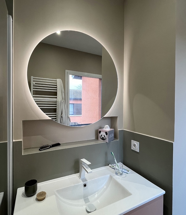 Salle de bain moderne : miroir rétroéclairé et lavabo Miroir rond rétroéclairé au-dessus d'un lavabo blanc moderne, avec niche contenant une boîte de mouchoirs panda et une brosse.
