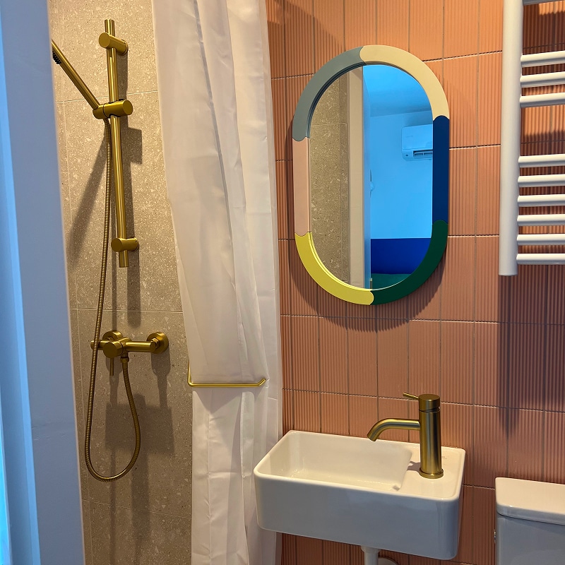 Salle de bain design : carrelage rose, laiton et miroir coloré Salle de bain moderne avec murs carrelés rose côtelé, robinetterie et porte-serviettes dorés, miroir ovale multicolore et lavabo blanc.